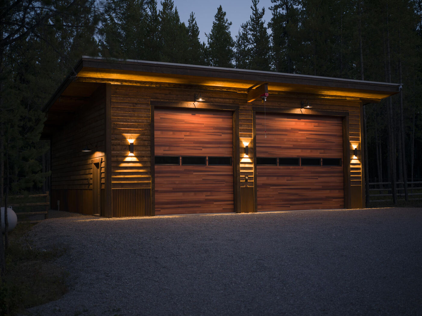 A two-bay timber garage at dusk, exterior lights washing down the cedar siding