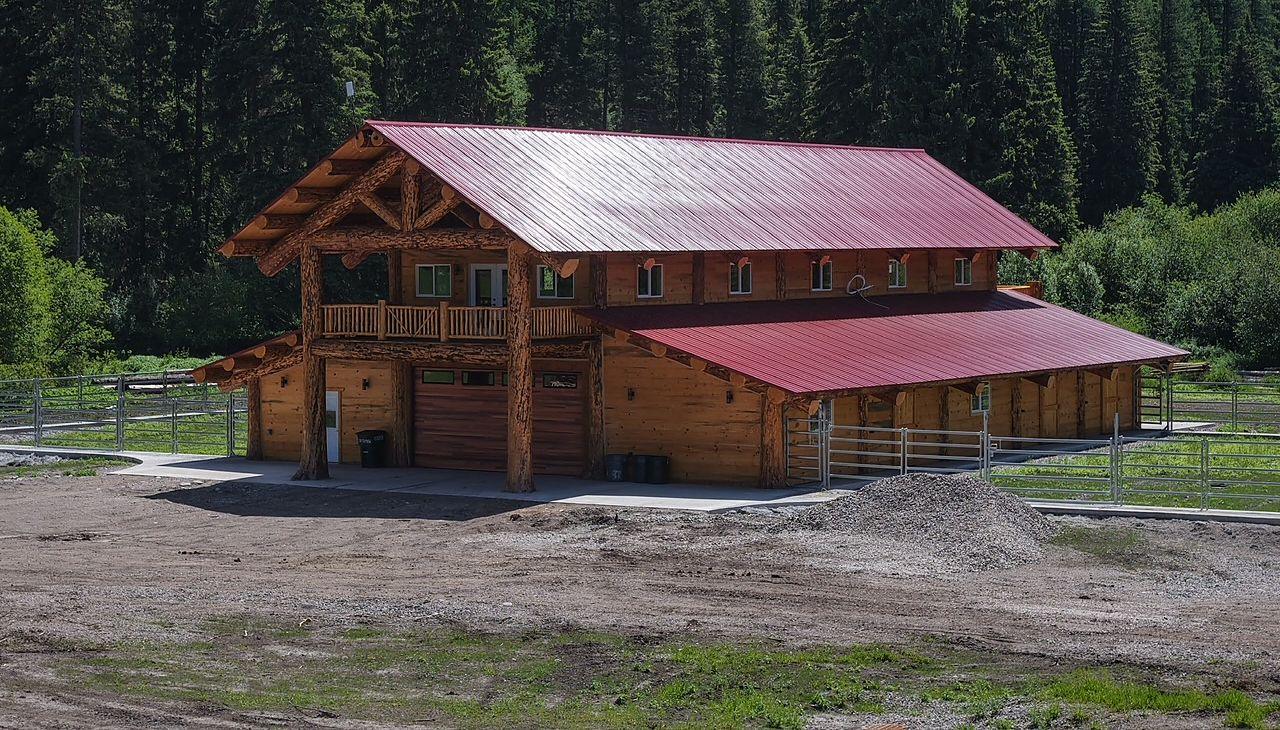 A hand-scribed log barn with a red metal roof in summer, forest behind and open ground in front